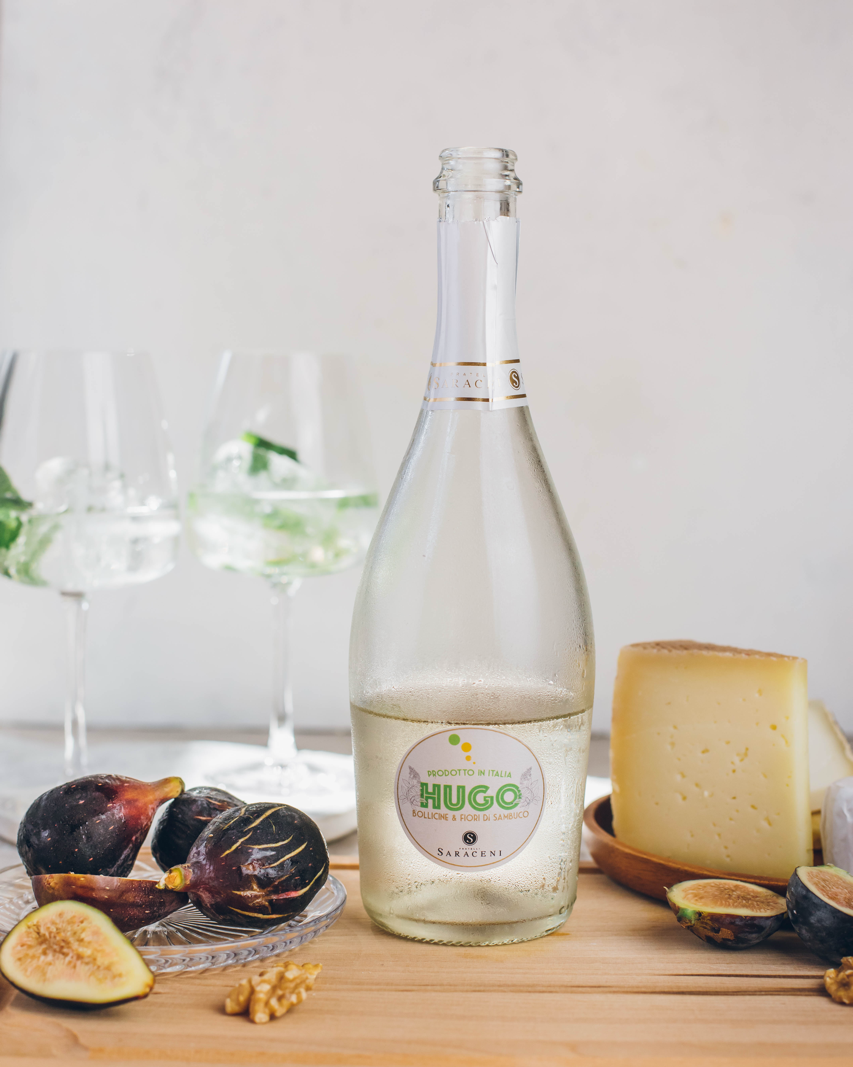 A bottle of Hugo sparkling wine with figs and cheese on a wooden board, with two wine glasses in the background.